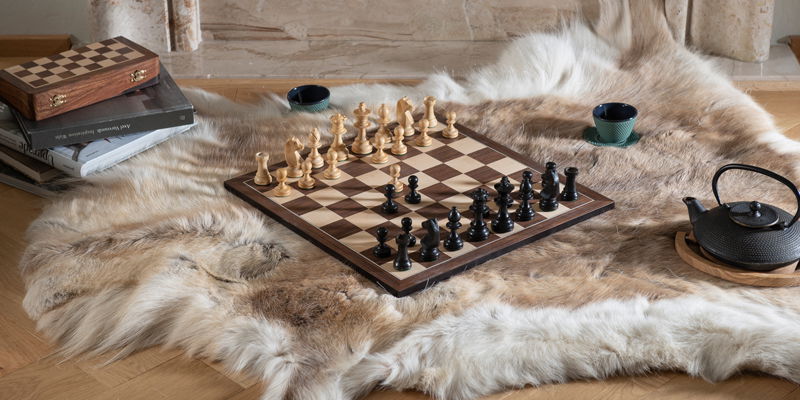 Schach - ein Spieklassiker für den Winter Schachset auf einem Fell am Boden mit Teekann und einem Stapel Büchern.